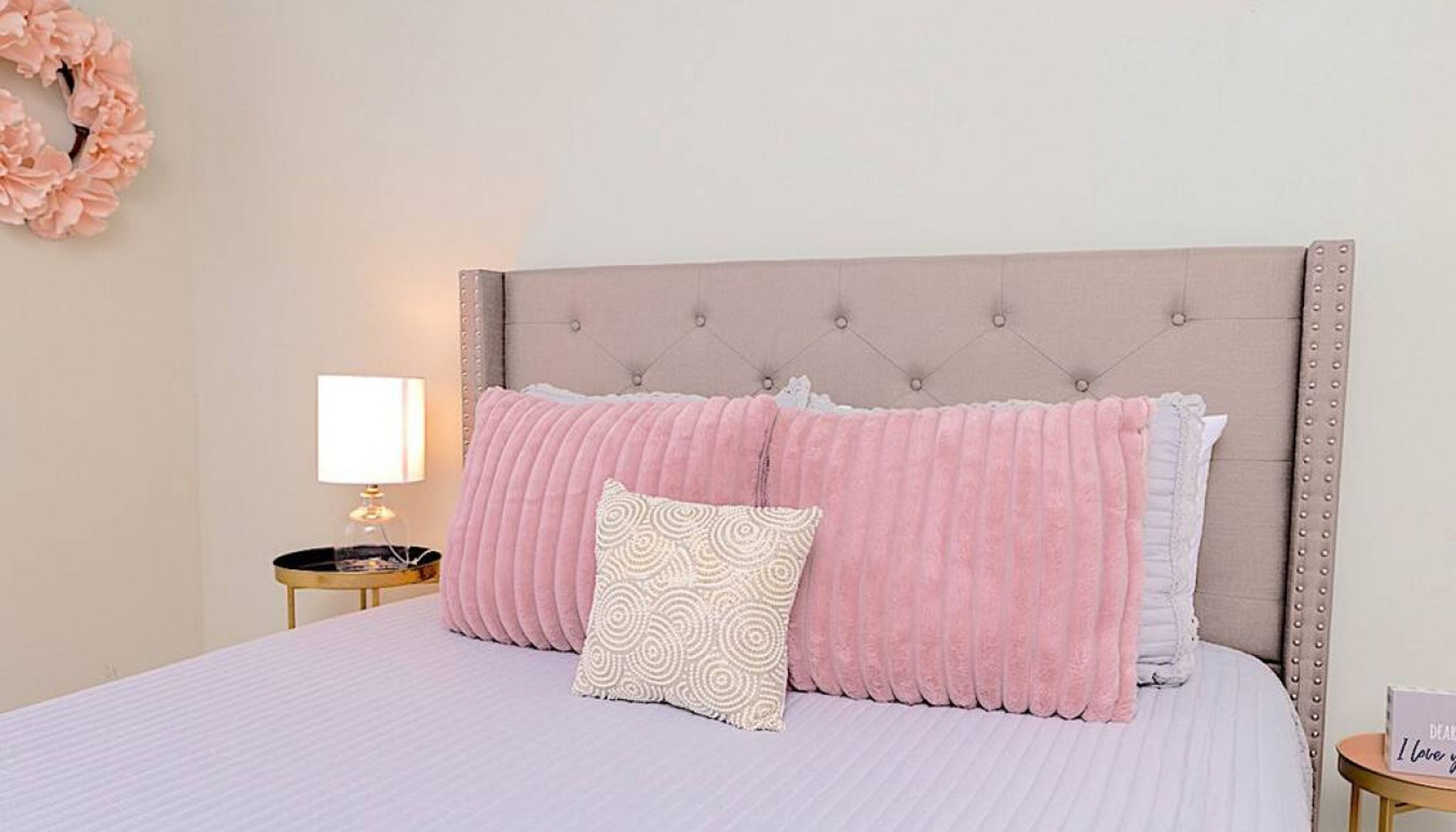 A neatly made bed with pink and white bedding, a tufted headboard, a bedside lamp, and a decorative wreath on the wall.
