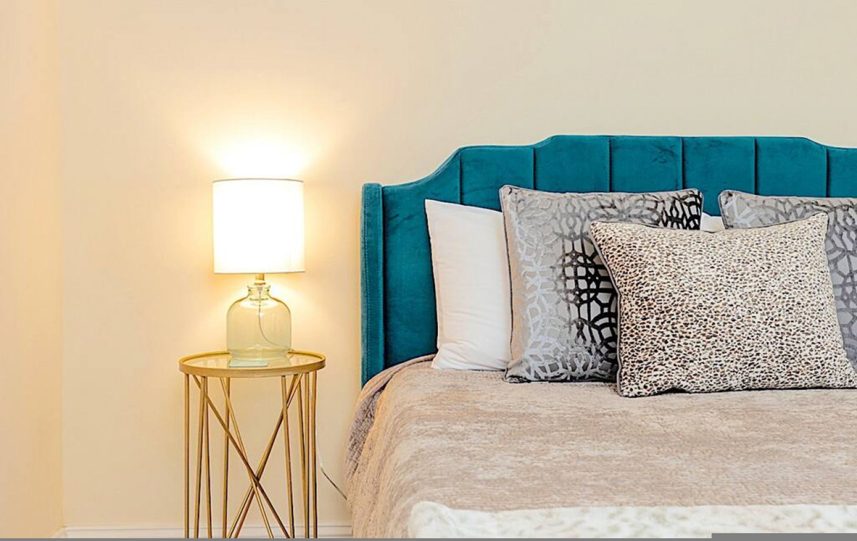 Un lit avec une tête de lit de couleur turquoise, une table de chevet ronde dorée surmontée d’une lampe en verre et des coussins à motifs.