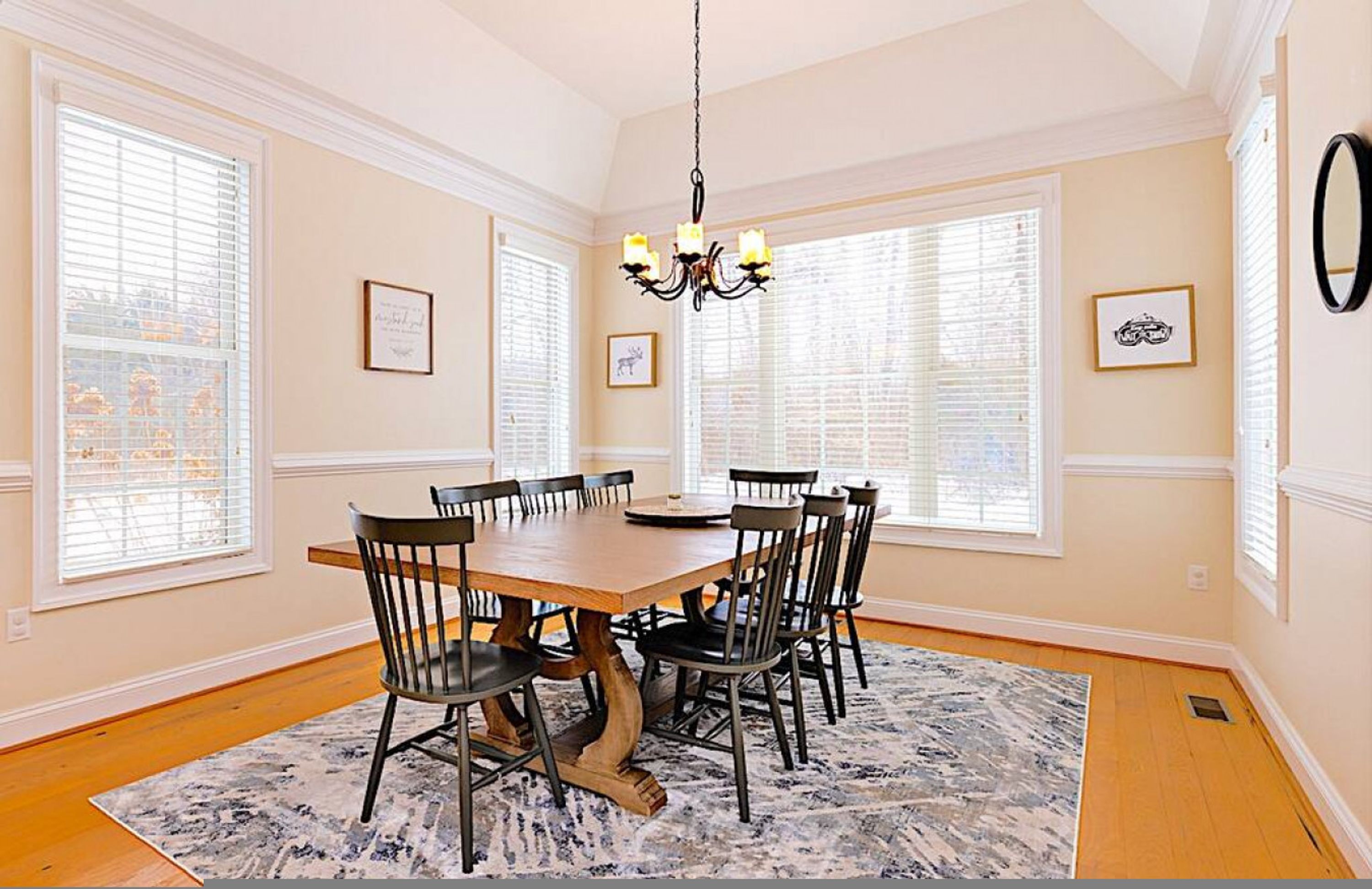 A dining room with a wooden table, six chairs, a chandelier, and large windows with blinds.