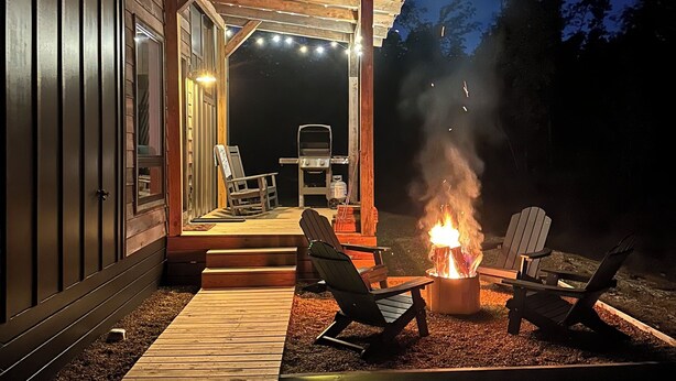 Een terras buiten met vuurkorf en lounge stoelen, dat uitkomt op een overdekte patio met lichtjes en een barbecue.