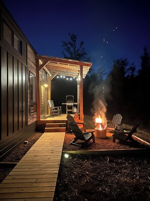 Una terraza exterior con un pozo de fuego y sillones de descanso, que da a un patio cubierto con luces colgantes y una parrilla.