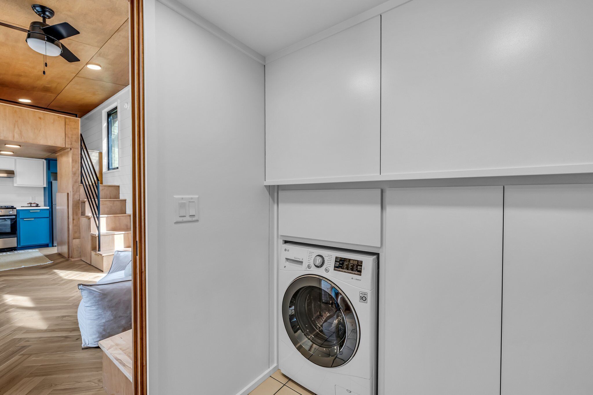 A laundry room with a front-loading washing machine, wooden flooring, and a staircase leading to a kitchen area.