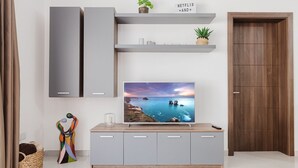 Un salon moderne avec un grand téléviseur à écran plat fixé sur un meuble gris, une porte en bois et une étagère ornée d’objets décoratifs.