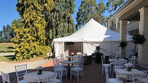 An outdoor patio with white tables and chairs, a white tent, and a building in the background.