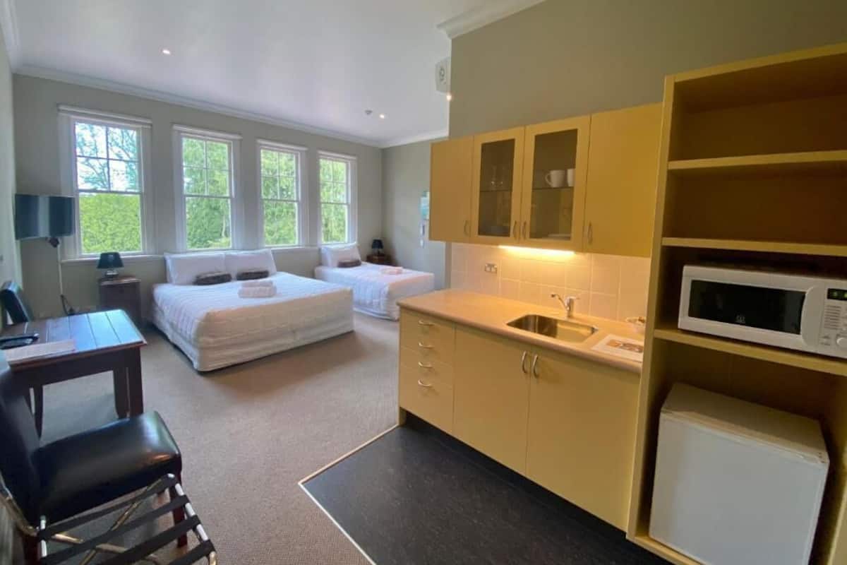 A compact hotel room with a bed, a desk, a chair, a kitchenette, and a large window.