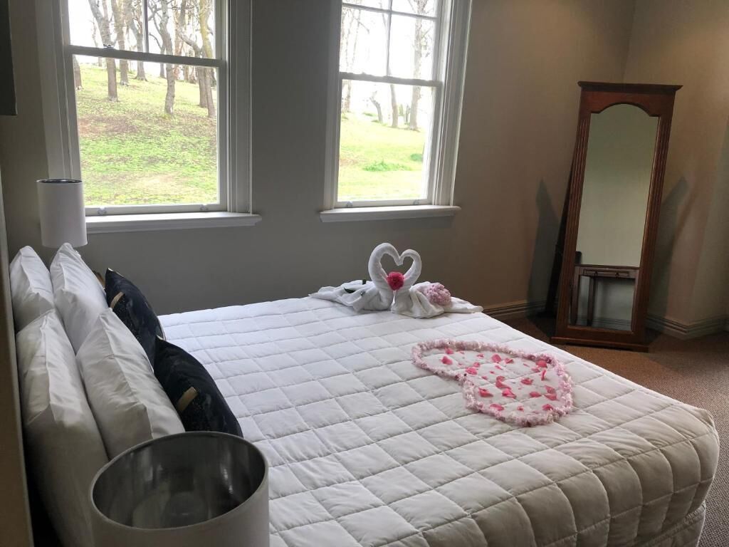 A neatly made bed with a white quilt, two pillows, and a heart-shaped decoration on the bed. There is a window with a view of trees and a wooden dresser with a mirror.