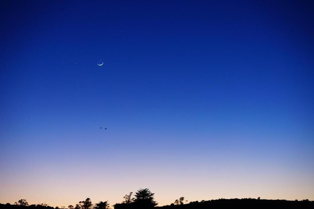 Cielo despejado con una luna creciente y algunos aviones a lo lejos.