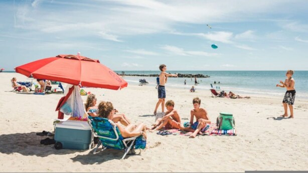 Escena de playa con gente relajándose bajo sombrillas, un enfriador y sillas de playa.