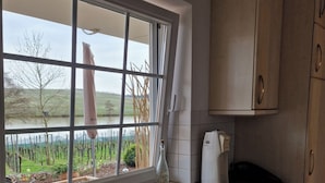 Une cuisine avec une fenêtre donnant sur un champ et des arbres, une bouteille posée sur le plan de travail et des armoires en bois.