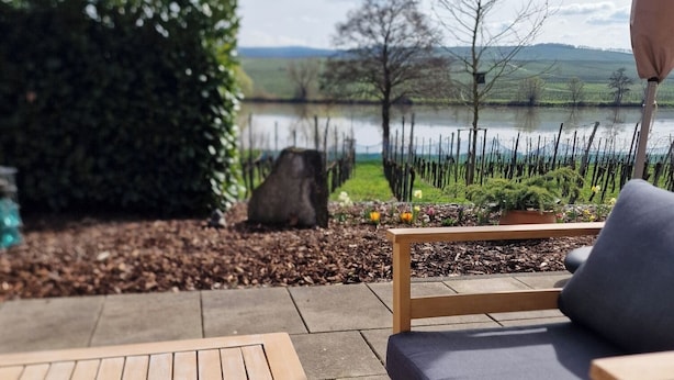 Une terrasse extérieure avec une table en bois et une chaise rembourrée, donnant sur un lac et un jardin agrémenté d’une clôture et d’arbres.
