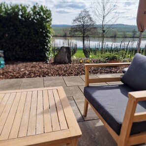 An outdoor patio with a wooden table and a cushioned chair, overlooking a lake and a garden with a fence and trees.