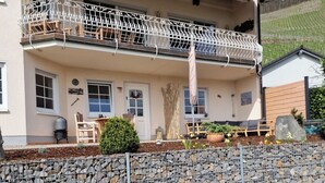 Une maison à deux étages, avec un mur de pierre à l’avant, un balcon doté de rampes en métal et un petit jardin agrémenté de plantes en pot et d’un banc en bois.
