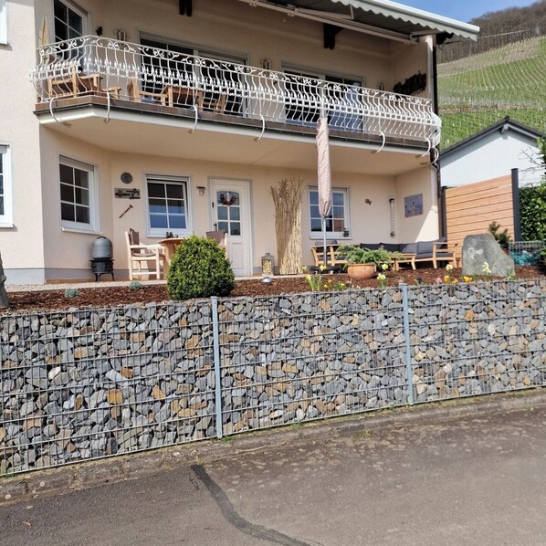 A two-story house with a stone wall in front, a balcony with metal railings, and a small garden area with potted plants and a wooden bench.