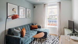 A modern living room with a blue sofa, a small round table, and a television. There are three framed artworks on the wall, a window with curtains, and a view of the outside balcony.