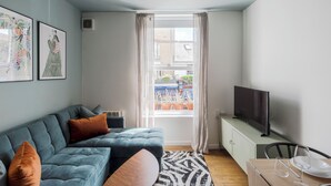 A modern living room with a sectional sofa, a flat-screen TV on a cabinet, and a view of the street through large windows.