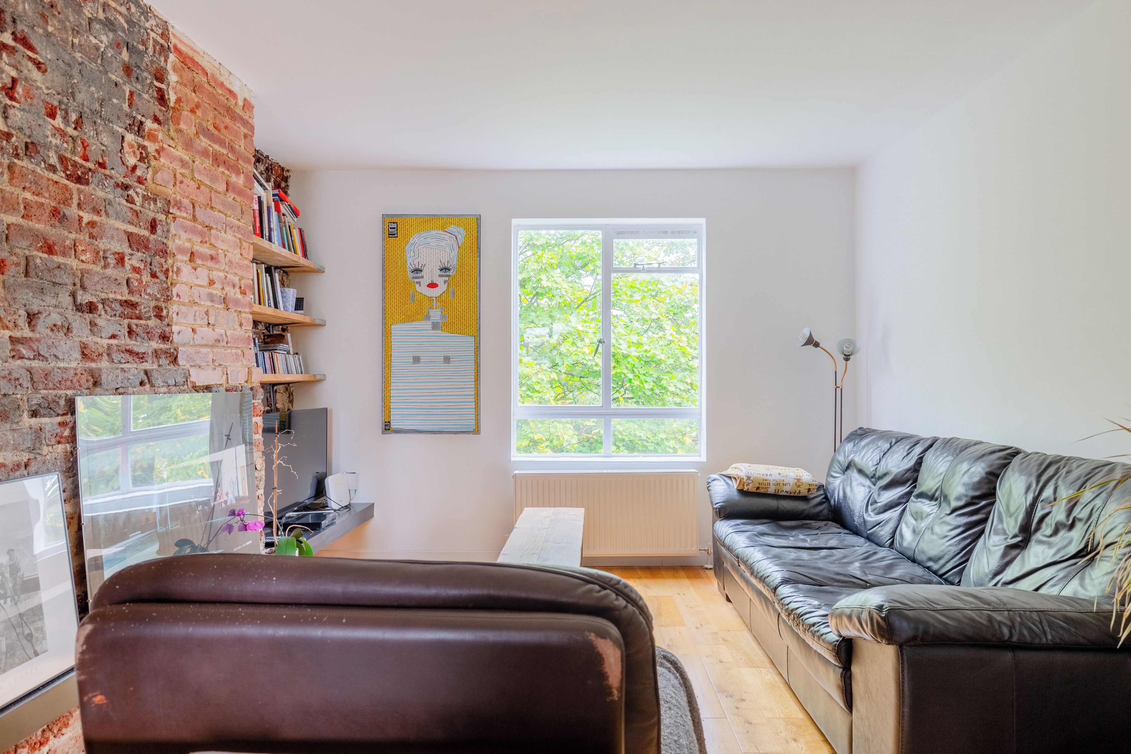 Een moderne woonkamer met een bakstenen muur, een zwarte leren bank, een boekenkast en een groot raam met uitzicht op groen.