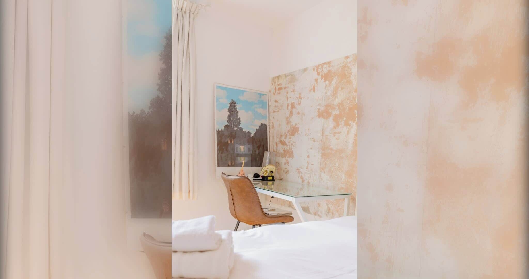 Une chambre d’hôtel avec un lit, une table en verre, une chaise, un tableau au mur et une fenêtre avec des rideaux.