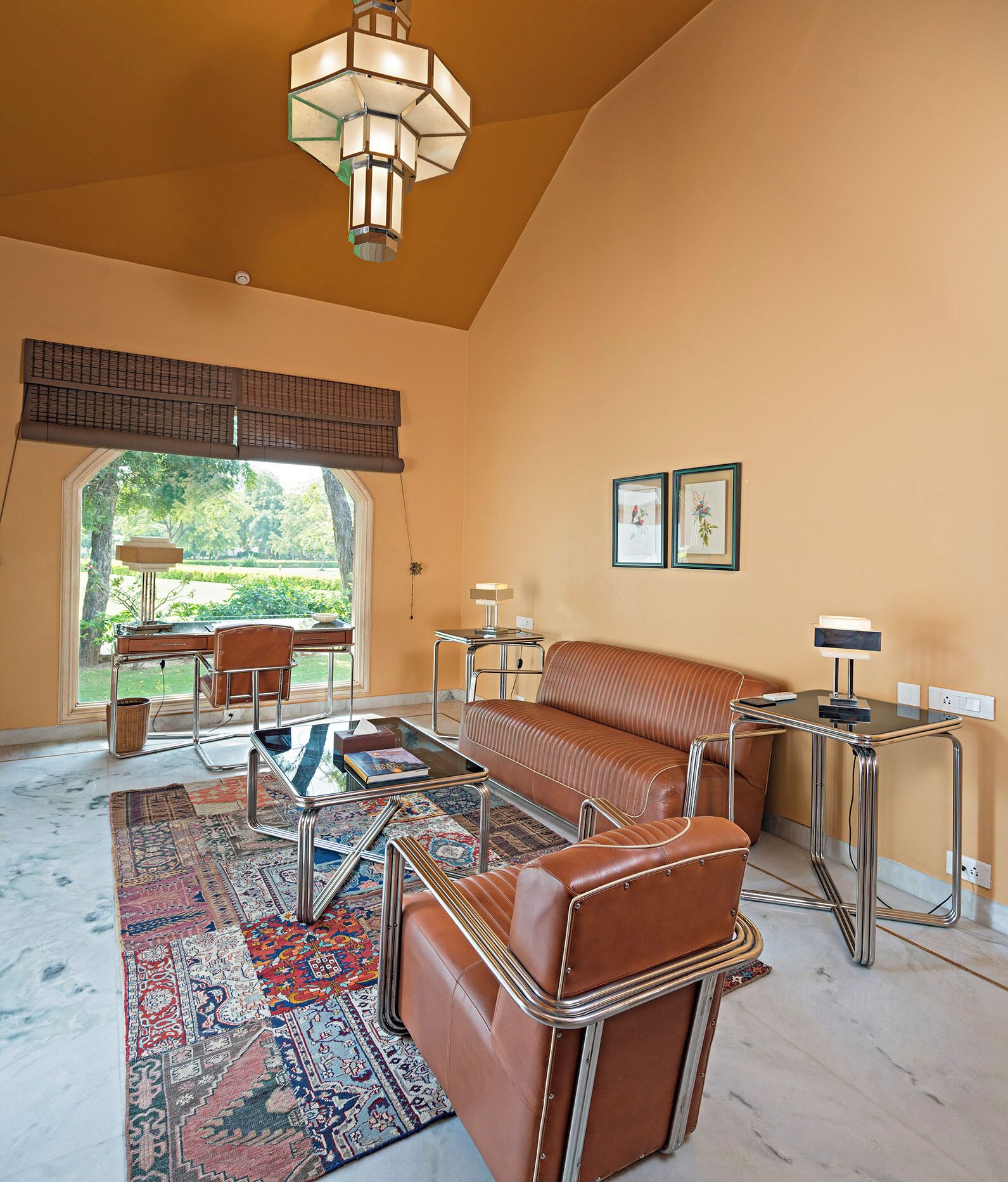 A spacious living room with a brown leather sofa, a glass coffee table, and a patterned rug. There is a large window with a view of greenery outside.