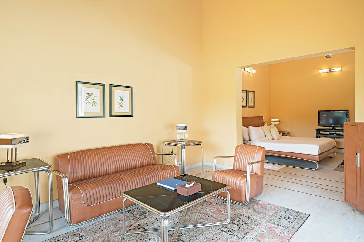 Habitación de hotel con sofá, sillón, mesa de centro y cama. Hay un televisor y una mesita de noche con lámpara.