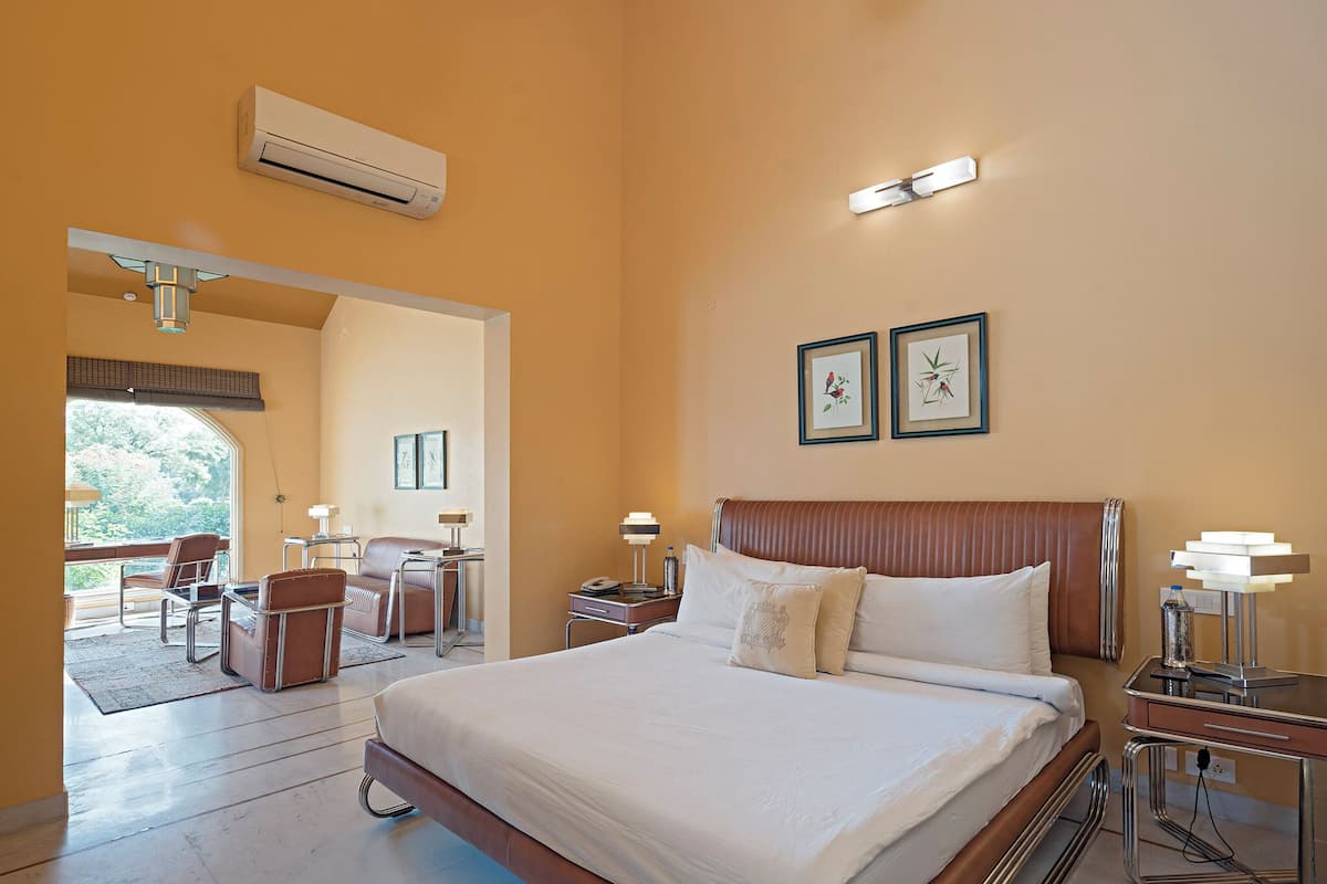 Una habitación de hotel con una cama grande, un escritorio con una silla, una mesita de noche con una lámpara y una ventana con vista a árboles.