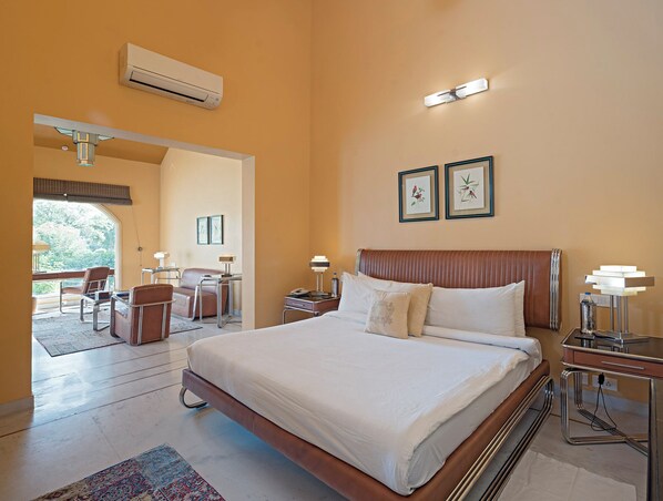 A hotel room with a large bed, a desk with a chair, a small table with a lamp, and a window with a view of trees. - Raj Niwas Palace (Dhaulpur)