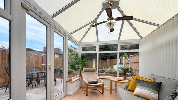 Une véranda dotée d’une porte vitrée donnant sur un patio, d’un ventilateur de plafond et d’un espace salon aménagé avec un canapé et des fauteuils.