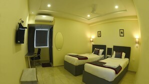 A hotel room with two beds, a desk, a chair, an air conditioning unit, and a small trash bin. - Seasons Stay (Vijayawada)