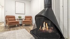 Een moderne woonkamer met een zwarte houtkachel, twee bruine leren fauteuils, een kamerbreed tapijt en een abstract kunstwerk in een lijst aan de muur.
