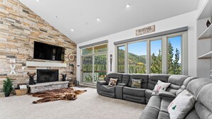 A living room with a stone fireplace, a gray sectional sofa, and a large window overlooking a forested area. - Spacious Condo Overlooking Keystone Lake w/ 2 Private Balconies & Fireplaces! (Keystone)