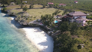Luftaufnahme einer Küstenimmobilie mit Strand, klarem Wasser und einem großen Haus mit Schwimmbad.