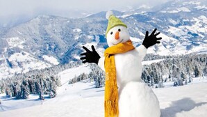 A snowman with a green hat and yellow scarf standing in a snowy landscape with mountains in the background.