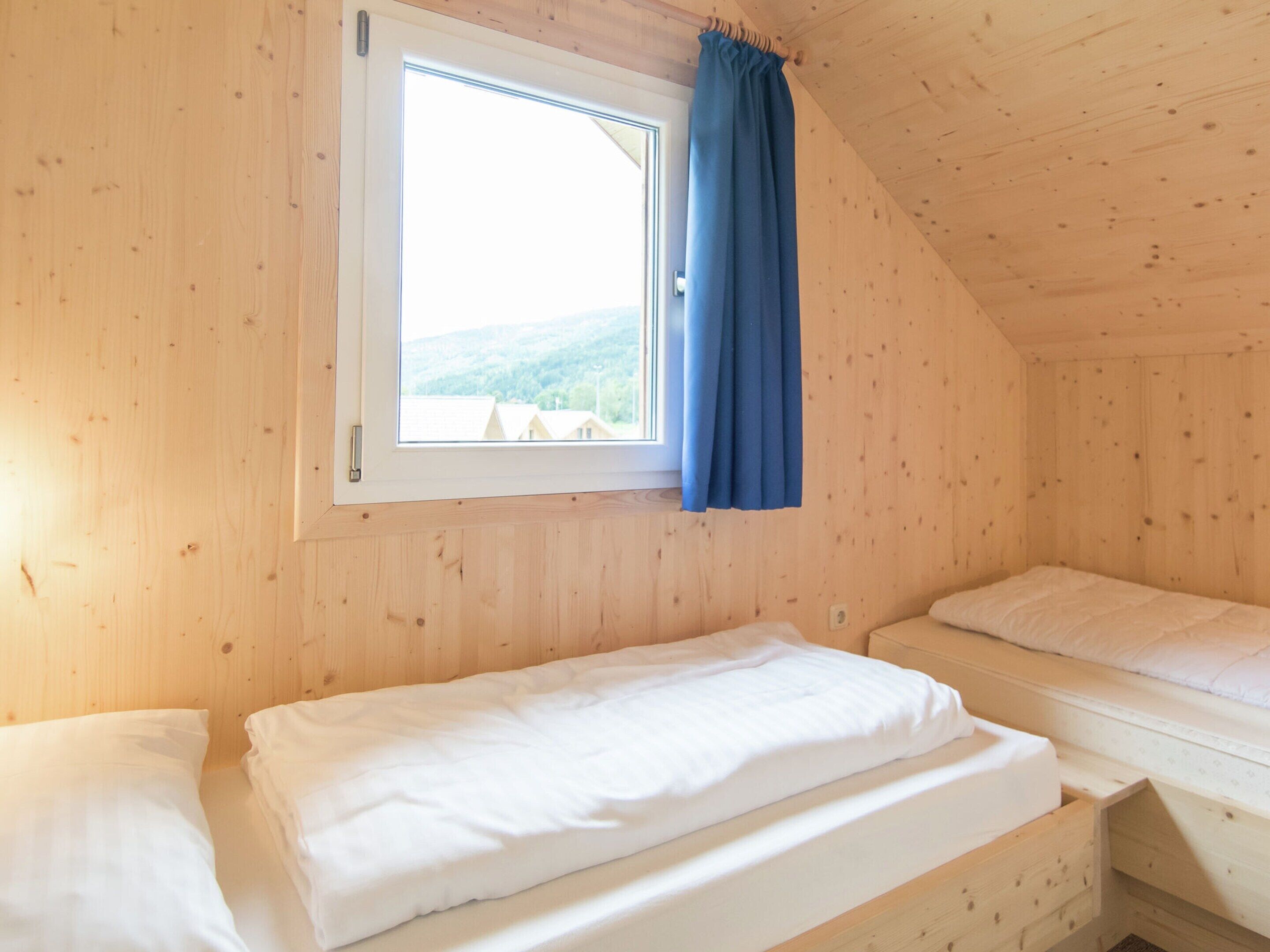 A wooden cabin room with two beds, a window with a curtain, and a view of a forested area.