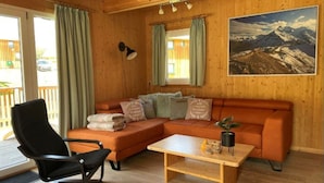 Interior acogedor de una cabaña con piso de madera, un sofá modular, un sillón negro, una mesita y un cuadro de un paisaje montañoso en la pared.