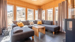 A modern living room with a sectional sofa, a small wooden table, and large windows offering a view of the outdoors. - Superior Chalet With Sauna (Sankt Georgen am Kreischberg)