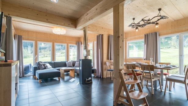 A spacious living area with a sofa, coffee table, and dining table set up. There are large windows providing natural light and views of the outdoors. - Superior Chalet With Sauna (Sankt Georgen am Kreischberg)
