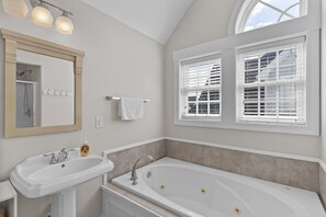 A bathroom with a sink, mirror, and bathtub. There are two windows with blinds, a towel rack, and a soap dispenser.