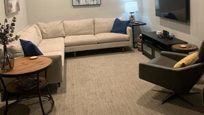 A modern living room with a beige sofa, a small round table, a side table with a lamp, a flat-screen TV mounted on the wall, and a decorative vase with branches. - Newer luxury cottage with hot tub; walking distance to town and beach (New Buffalo)