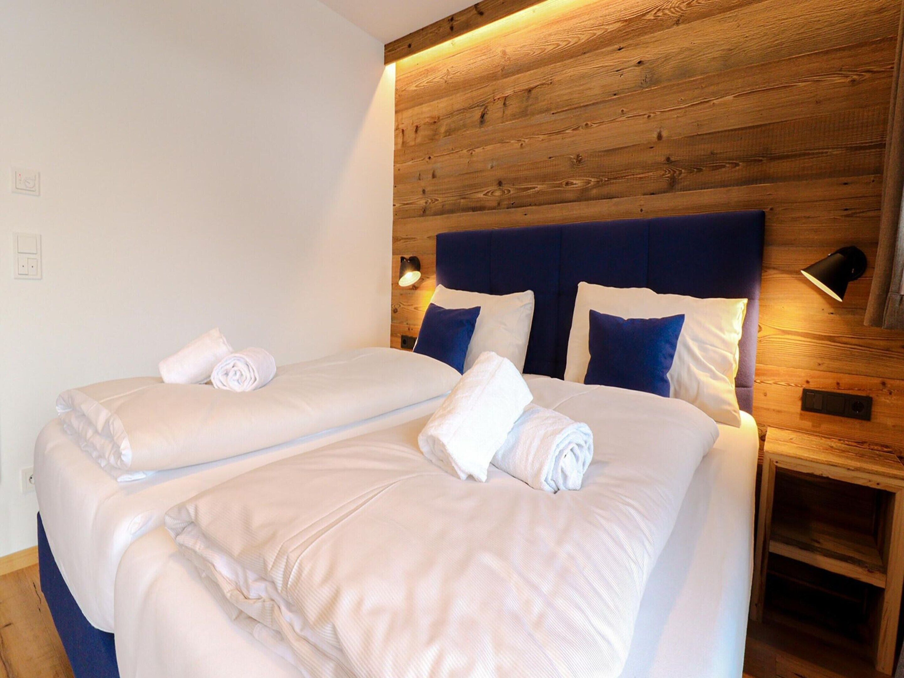 Cama de casal com lençóis brancos e almofadas azuis, cabeceira de madeira e mesa de cabeceira com abajur.