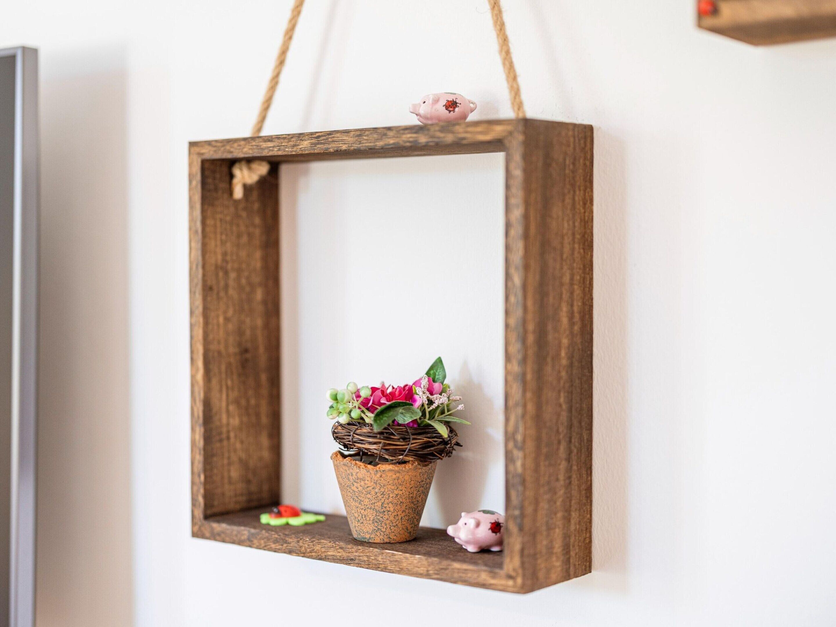A rustic wooden frame with a woven basket containing pink flowers and green foliage, hanging on a white wall.