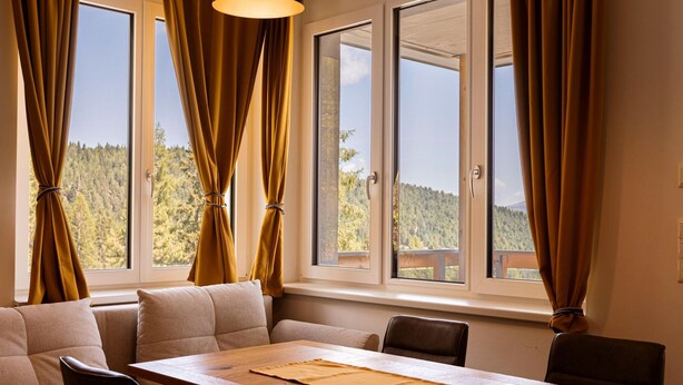 A room with a wooden table, beige sofa, and curtains tied back to reveal a mountain view. - Nice Apartment With Balcony or Terrace (Stadl-Predlitz)