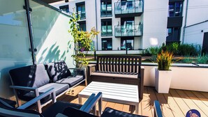 Un espacio al aire libre con sofás, sillas y una mesa, rodeado de plantas en macetas y un edificio moderno al fondo.