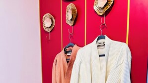 Una pared roja con tres abrigos colgados en ganchos, un banco tapizado y sombreros decorativos en la pared.