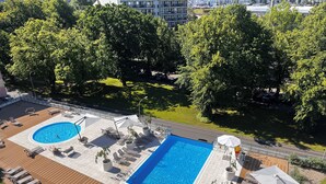 Una piscina en la azotea con dos zonas separadas, rodeada de sillones de descanso y sombrillas, con vistas a árboles y edificios.