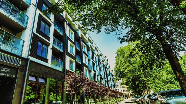 Un edificio de varios pisos con balcones, una calle bordeada de coches aparcados y árboles.