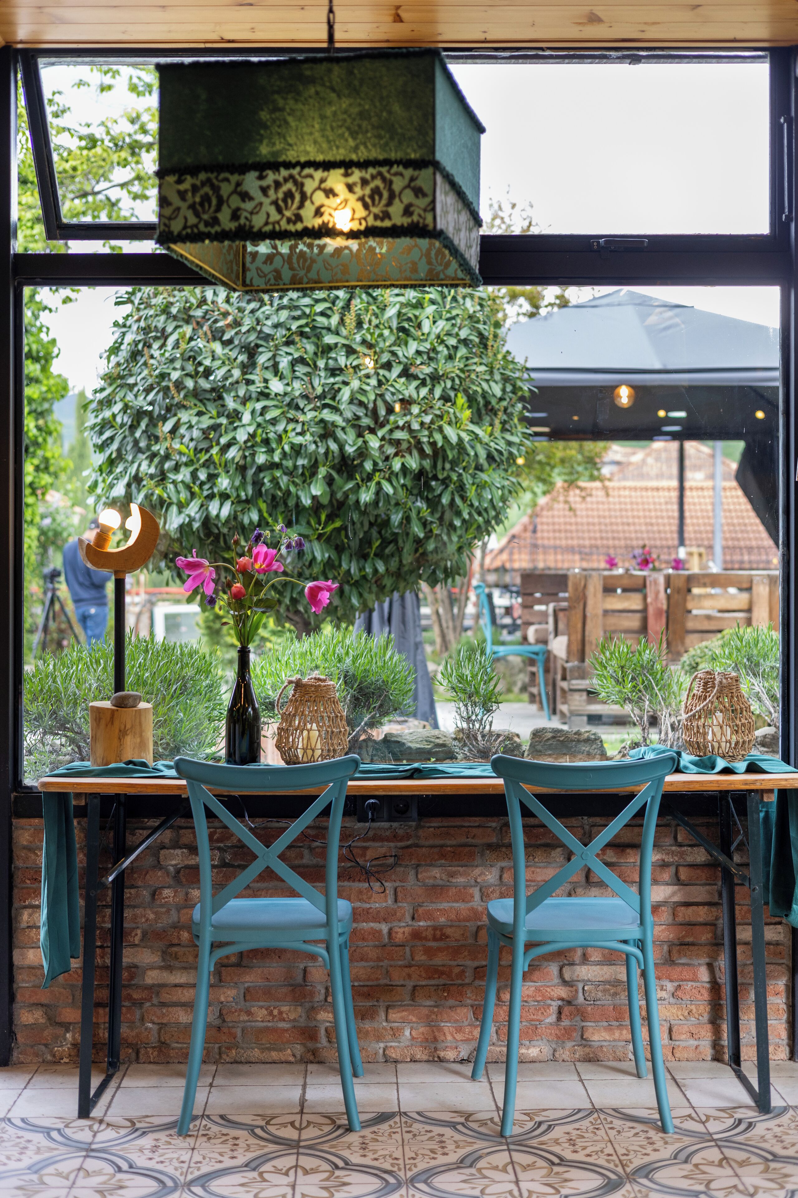 A restaurant with a brick wall, two teal chairs, a table with a vase of flowers, and a view of a tree and buildings outside.