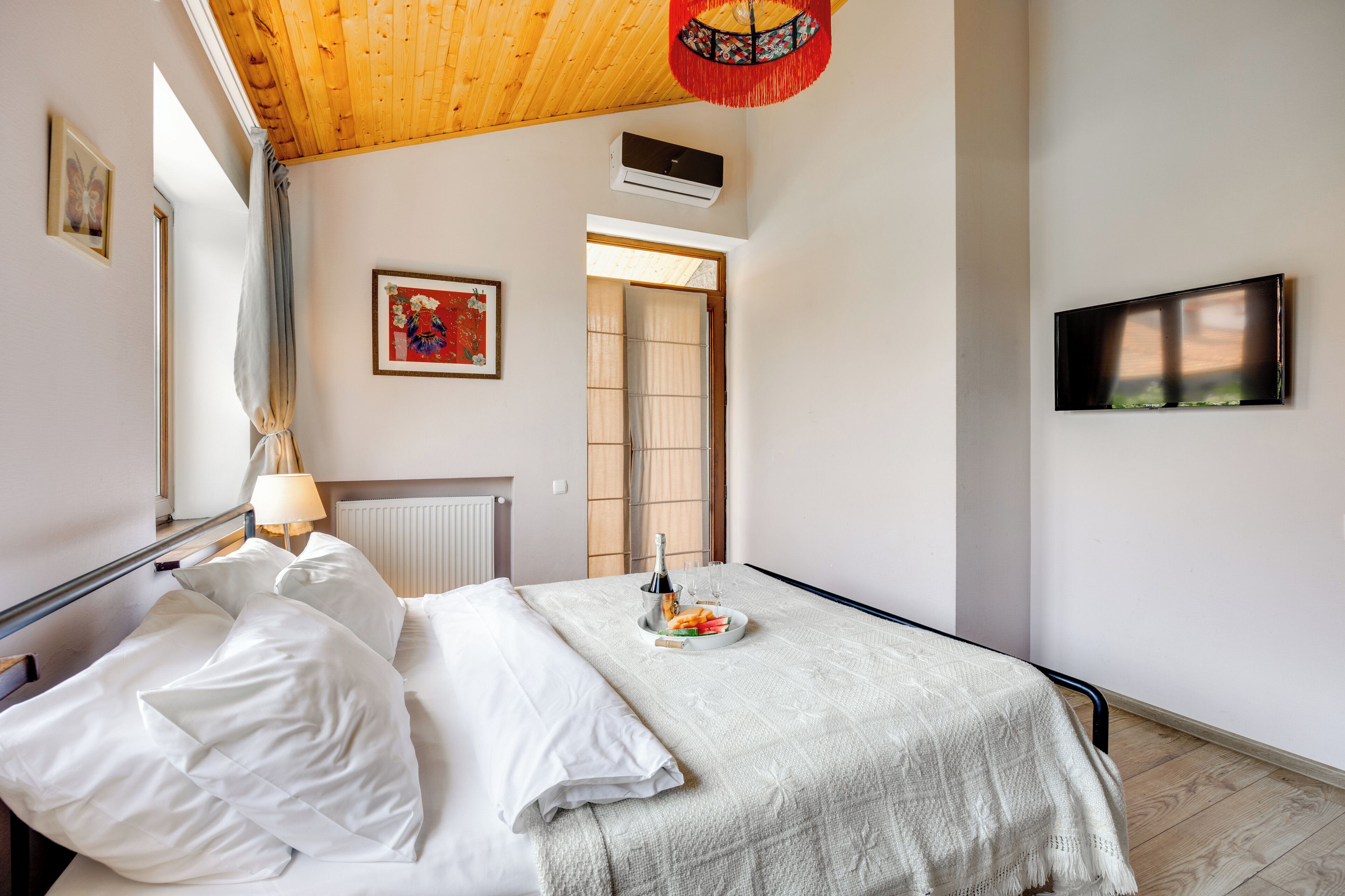 A hotel room with a wooden ceiling, a bed with white bedding, a small table with a bottle and glasses, a TV on the wall, and a window with a view.