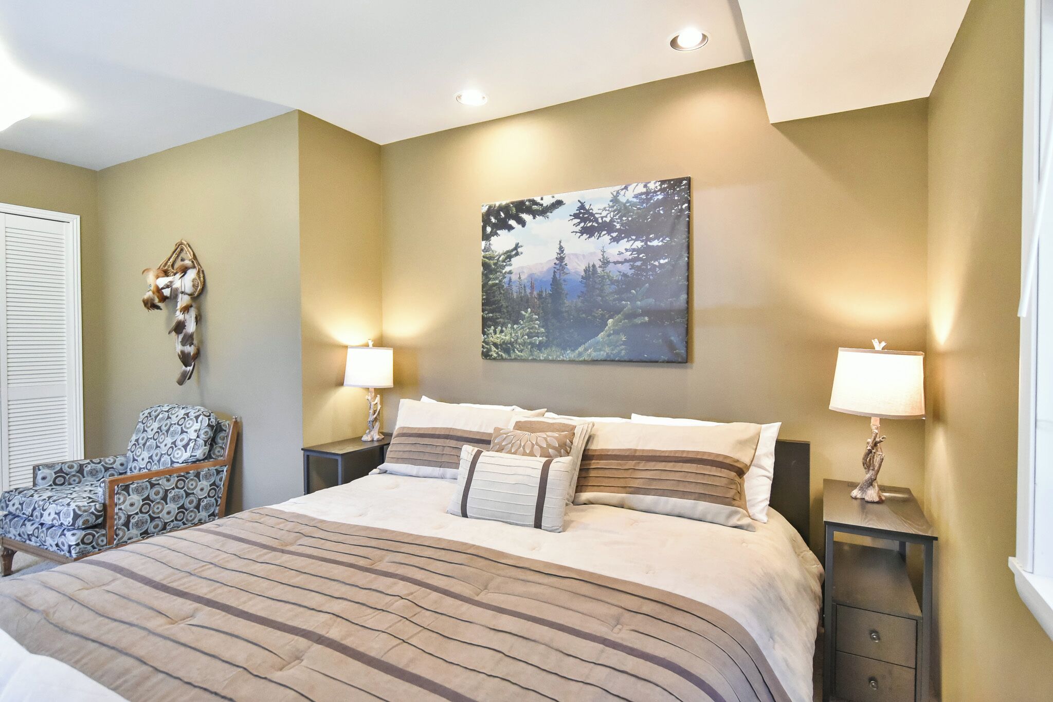 Une chambre à coucher avec un grand lit, deux tables de chevet, une chaise, un élément décoratif fixé au mur et un tableau.
