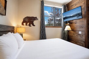 A hotel room with a bed, a mounted television, a wall-mounted lamp, a window with curtains, and a bear silhouette decoration.