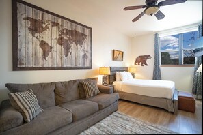 Un dormitorio con cama, sofá, cuadro de un mapa mundial, ventilador de techo y ventana con vista al cielo.
