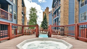 A hot tub surrounded by a wooden deck, with a pool in the background and a tree. - River Mountain Lodge Aspen Trail Condo: Ski In, Downtown Breck (Breckenridge)